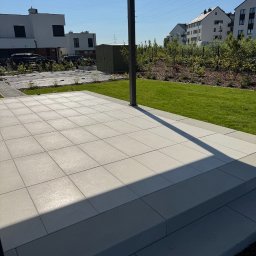 GreenPolis Sp. z o.o. - Nowoczesny taras z jasnoszarej kostki brukowej, schody tarasowe, równo przystrzyżony trawnik i zadbane nasadzenia w tle, widok z zadaszenia.