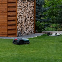 GreenPolis Sp. z o.o. - Zadbany ogród z robotem koszącym na trawniku, drewnianą wiatą na drewno i dekoracyjnym ogrodzeniem. Widok na zieloną przestrzeń z elementami architektury ogrodowej.