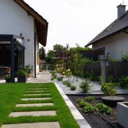GreenPolis Sp. z o.o. - Nowoczesny ogród z trawnikiem, ścieżką z płyt, rabatami z białym grysem i roślinami. Widok na dom z pergolą i meblami ogrodowymi w tle.