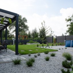GreenPolis Sp. z o.o. - Nowoczesny ogród z szarą kostką, żwirem, równo przystrzyżonym trawnikiem i basenem w tle. Pergola z meblami ogrodowymi po lewej stronie kadru.