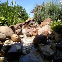 Ogr&oacute;d skalny z kaskadą wodną, otoczony zielenią i roślinami, z dekoracyjną lampą solarną w tle.