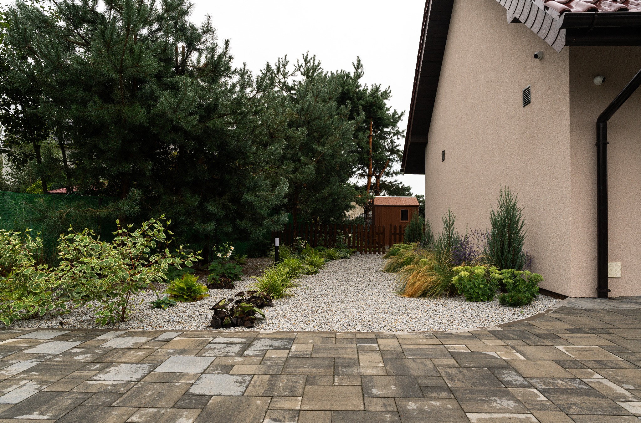 Ogród z ścieżką z drobnego żwiru, obrzeża obsadzone roślinami ozdobnymi, widok z kostki brukowej w kierunku drewnianej furtki i budynku.