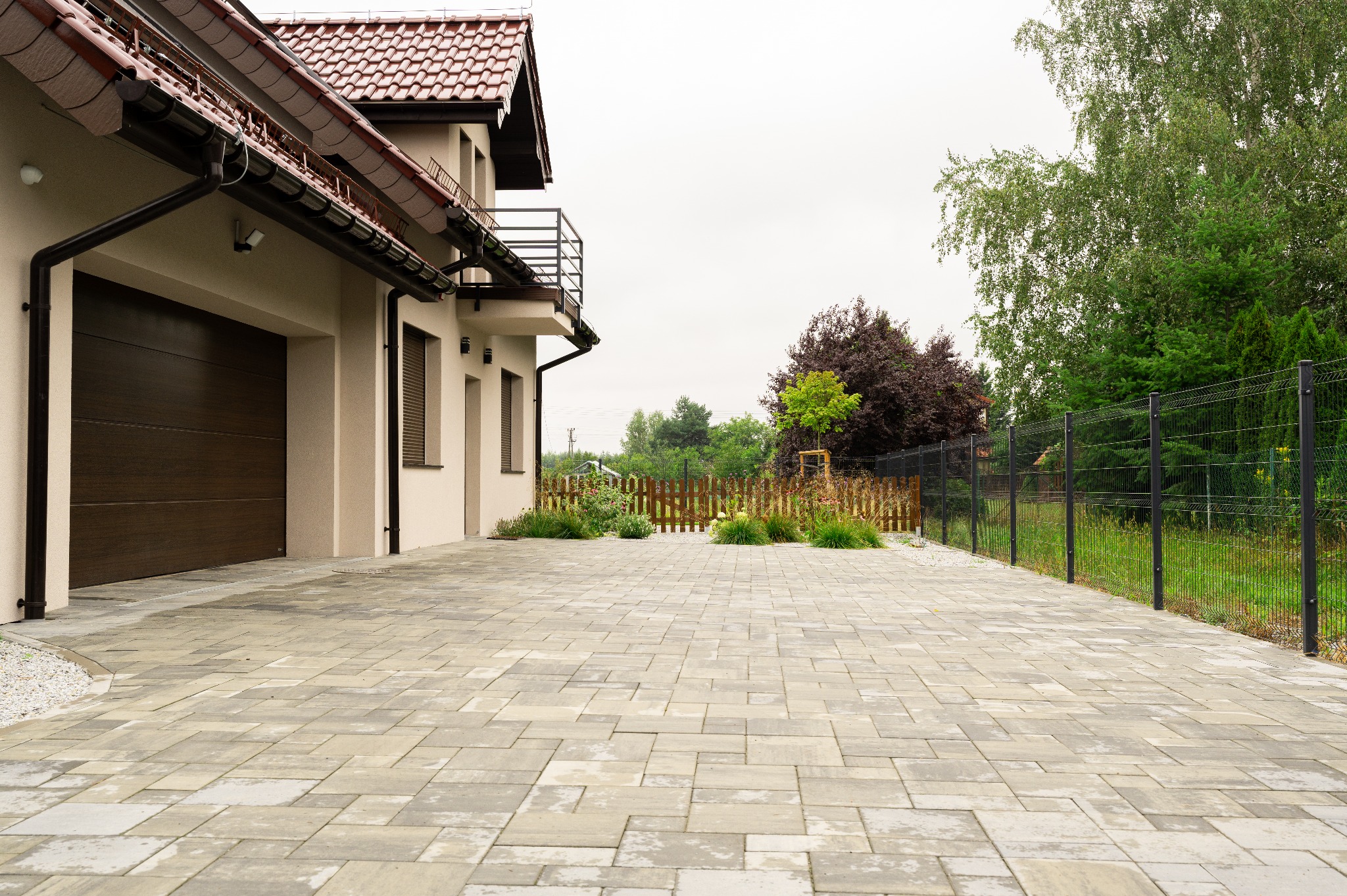 Ułożona kostka brukowa na podjeździe domu jednorodzinnego. Widoczny fragment budynku z garażem i ogrodzenie z roślinnością w tle. Szara kostka w różnych odcieniach.