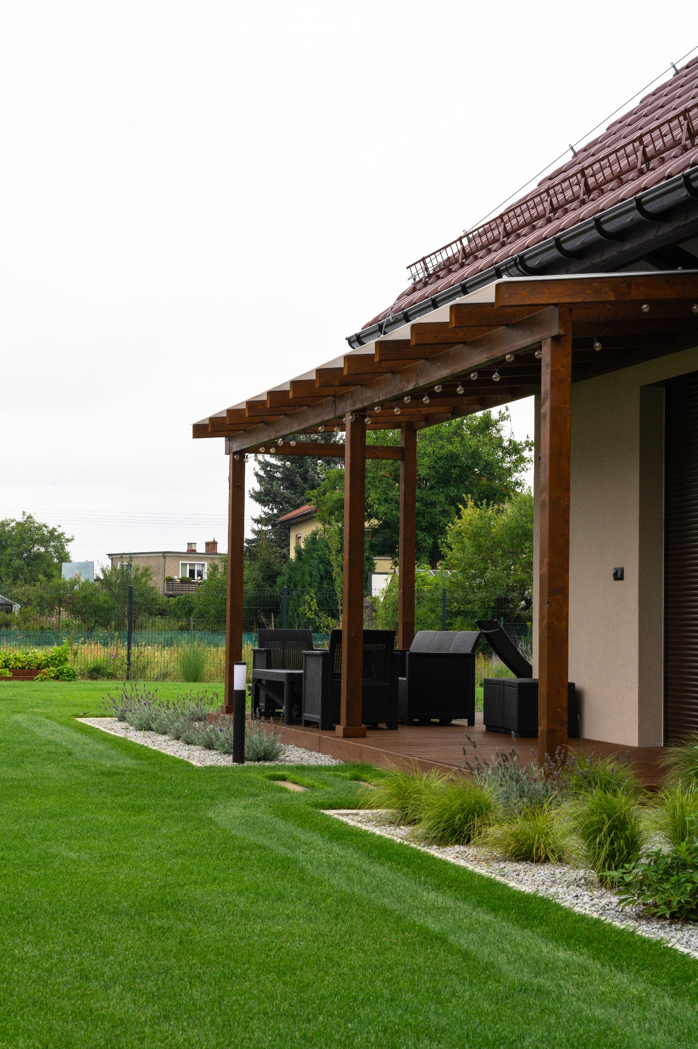 Pergola z oświetleniem nad tarasem, równo przystrzyżony trawnik z obrzeżami z kamyków i roślin. Widok na zadbany ogród i domy w tle.