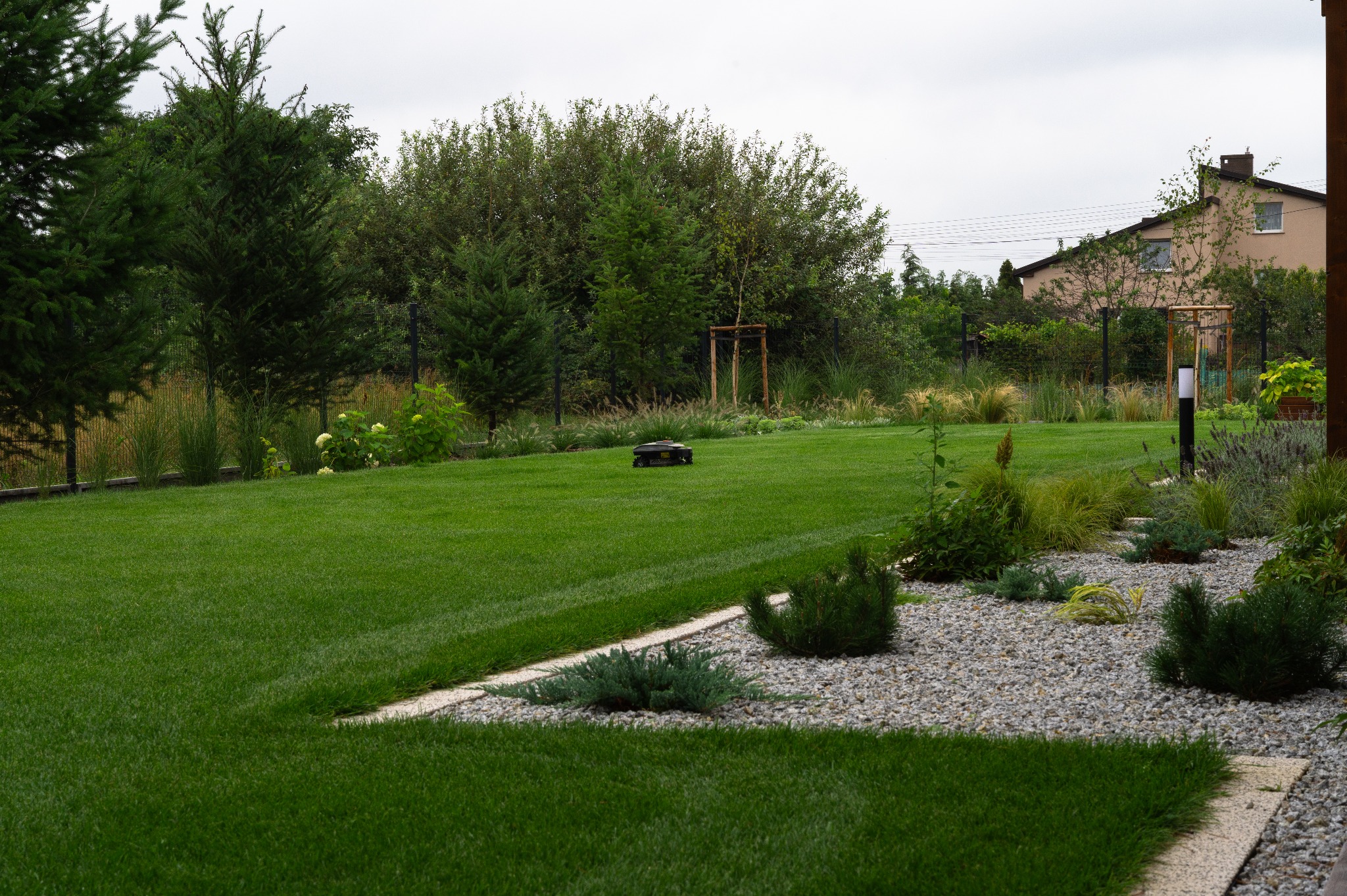 Zadbany trawnik z robotem koszącym na tle zieleni i domu. Granica trawnika wyłożona kamieniami. Widok na ogród w pochmurny dzień.