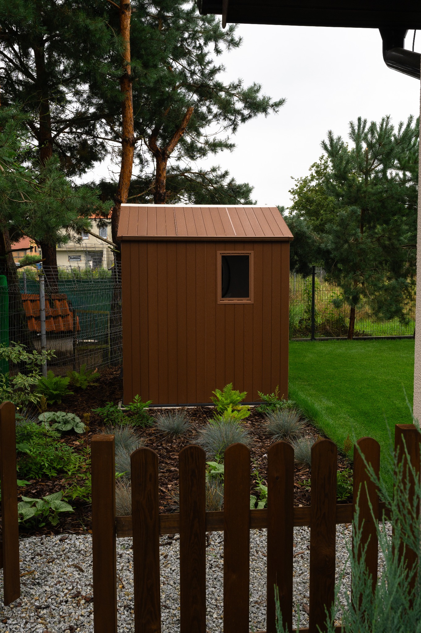Brązowy domek narzędziowy z oknem, otoczony rabatą z roślin i korą, za niskim drewnianym płotkiem. W tle drzewa i trawnik.