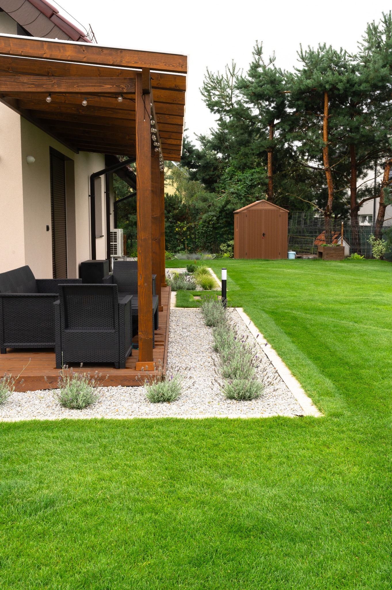 Nowoczesny ogród z równo przystrzyżonym trawnikiem, drewnianą pergolą z meblami ogrodowymi i ścieżką z białego żwiru, obok domu z beżową elewacją.