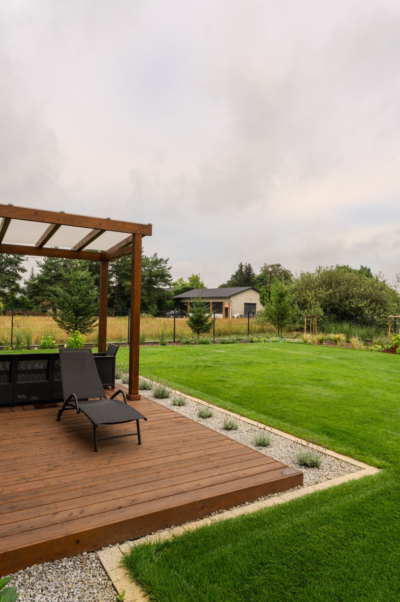 Pergola z drewnianym tarasem i leżakiem na tle zadbanego trawnika. Elementy małej architektury ogrodowej, obrzeża z kamienia. W tle budynek i drzewa.