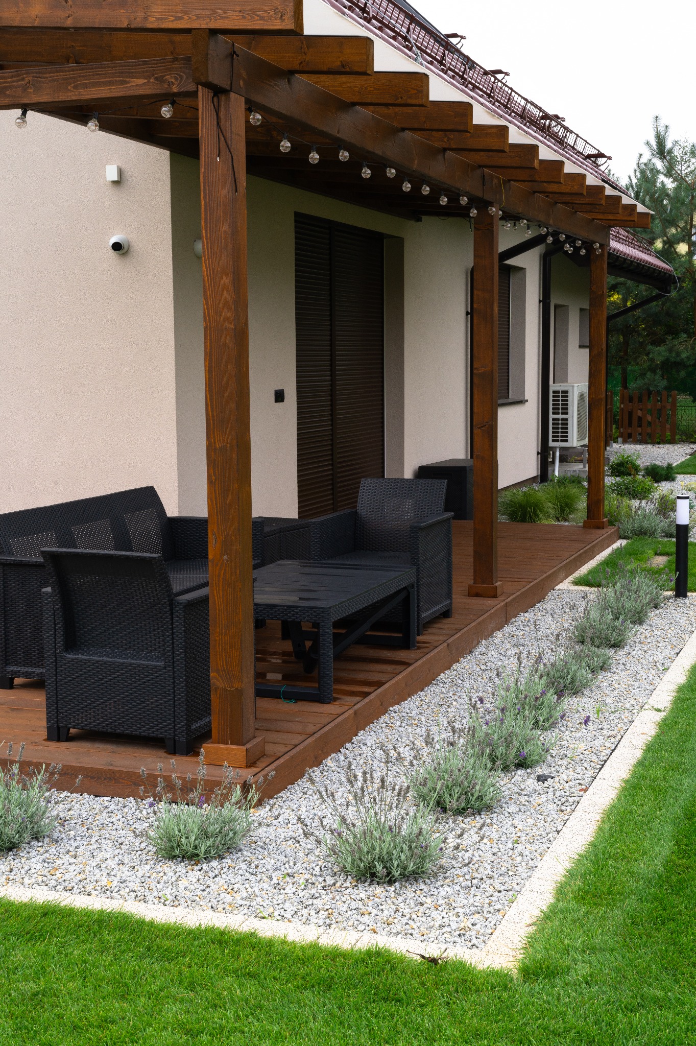 Zadaszony taras z drewnianą pergolą, oświetleniem i meblami ogrodowymi. Obok rabata z kamyków i lawendy, a dalej trawnik. Nowoczesny design.