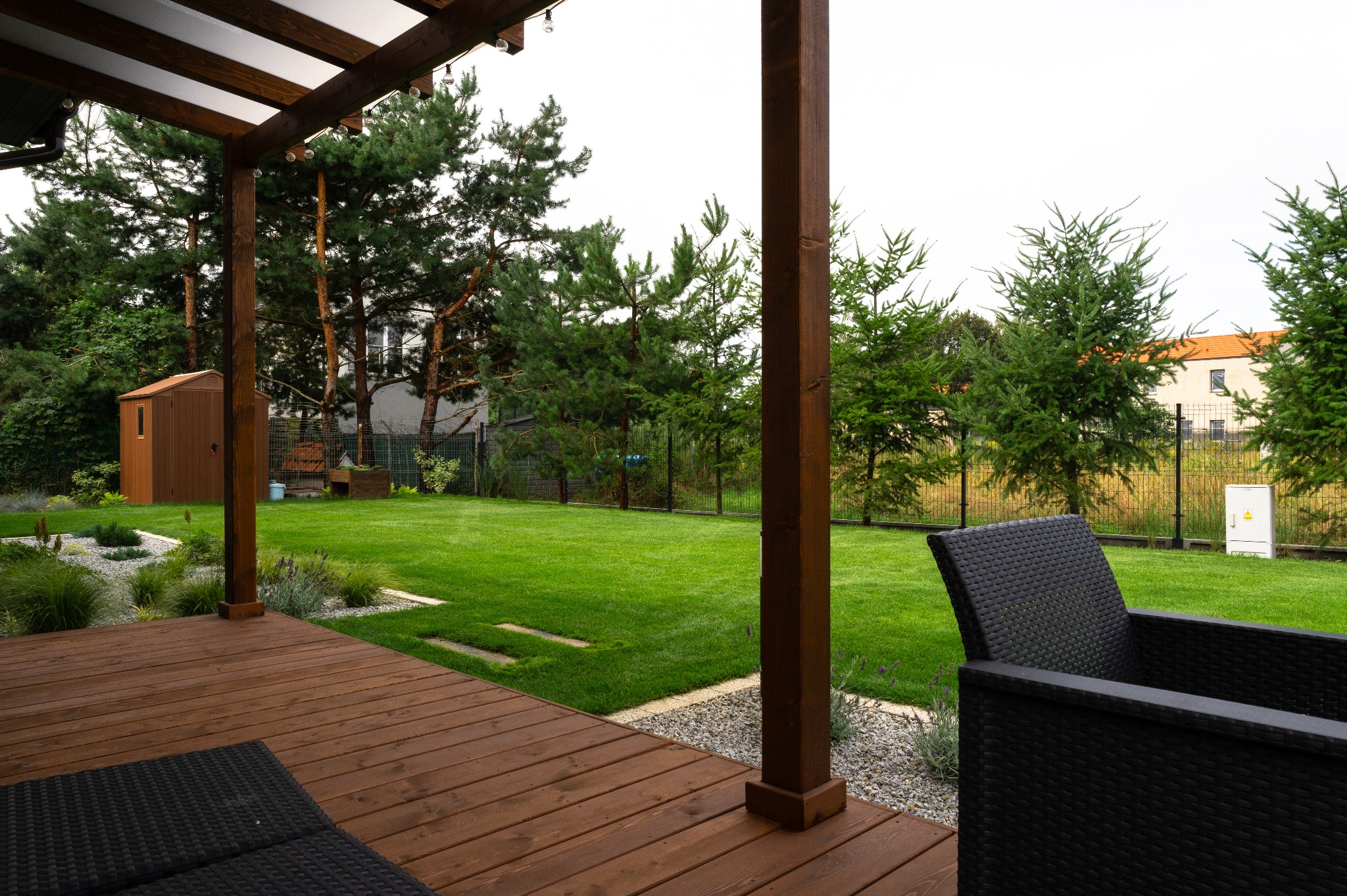Zadaszony taras z drewnianą podłogą i meblami ogrodowymi z widokiem na zadbany trawnik, drzewa iglaste i mały domek narzędziowy w tle.