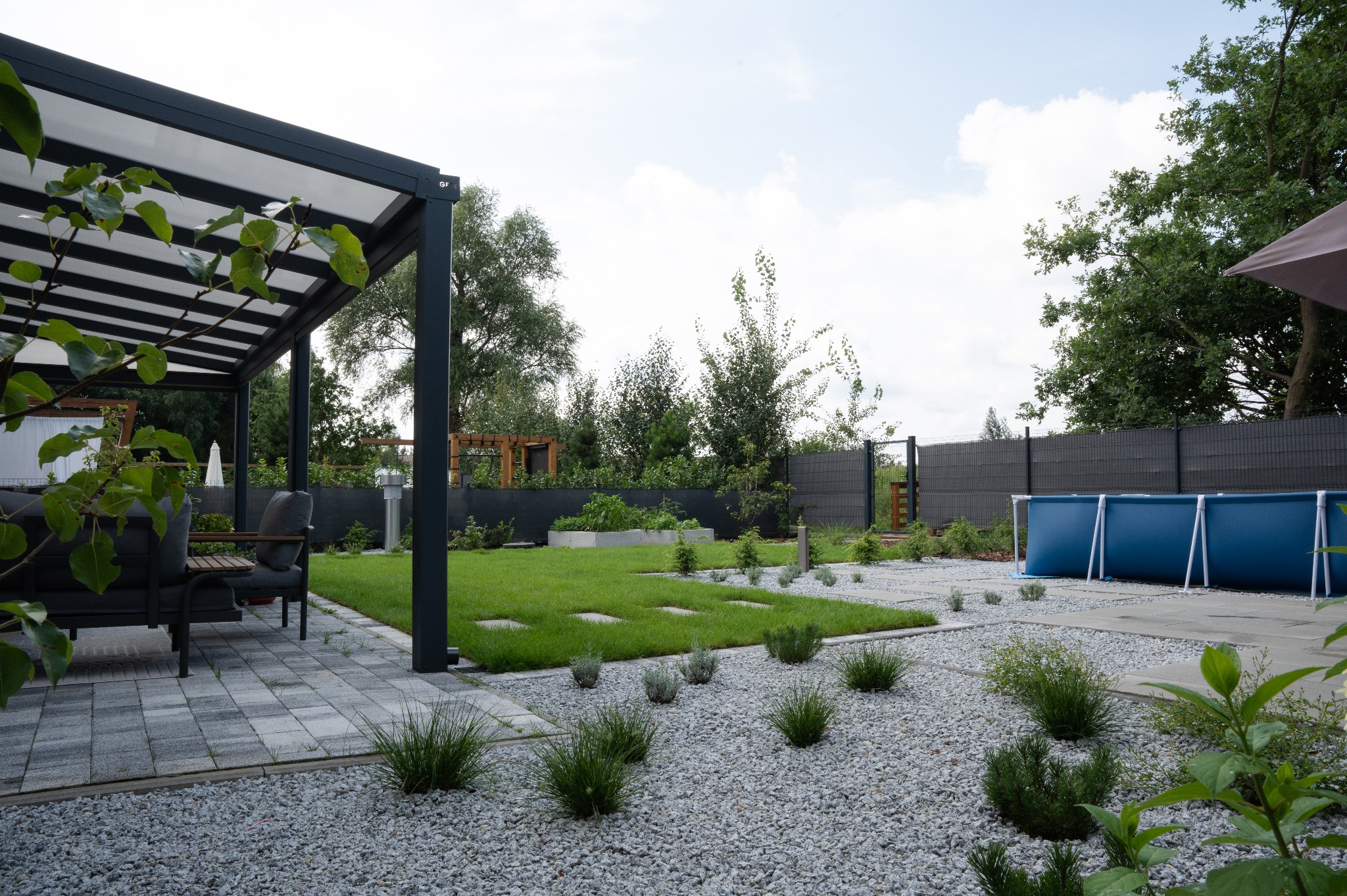 Nowoczesny ogród z szarą kostką, żwirem, równo przystrzyżonym trawnikiem i basenem w tle. Pergola z meblami ogrodowymi po lewej stronie kadru.
