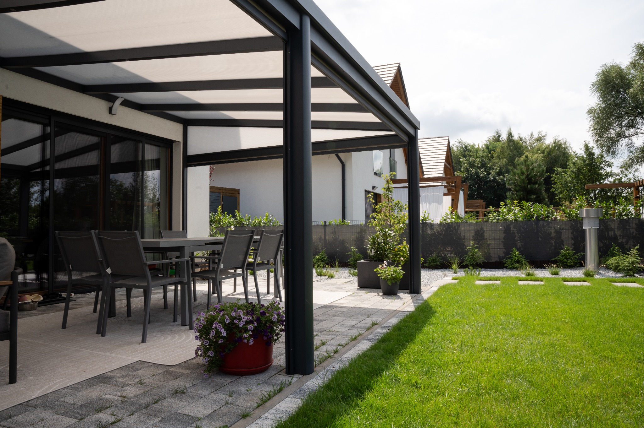 Nowoczesna pergola z zadaszeniem nad tarasem, szare meble ogrodowe, donica z kwiatami, zadbany trawnik i kostka brukowa. Elementy architektury krajobrazu.