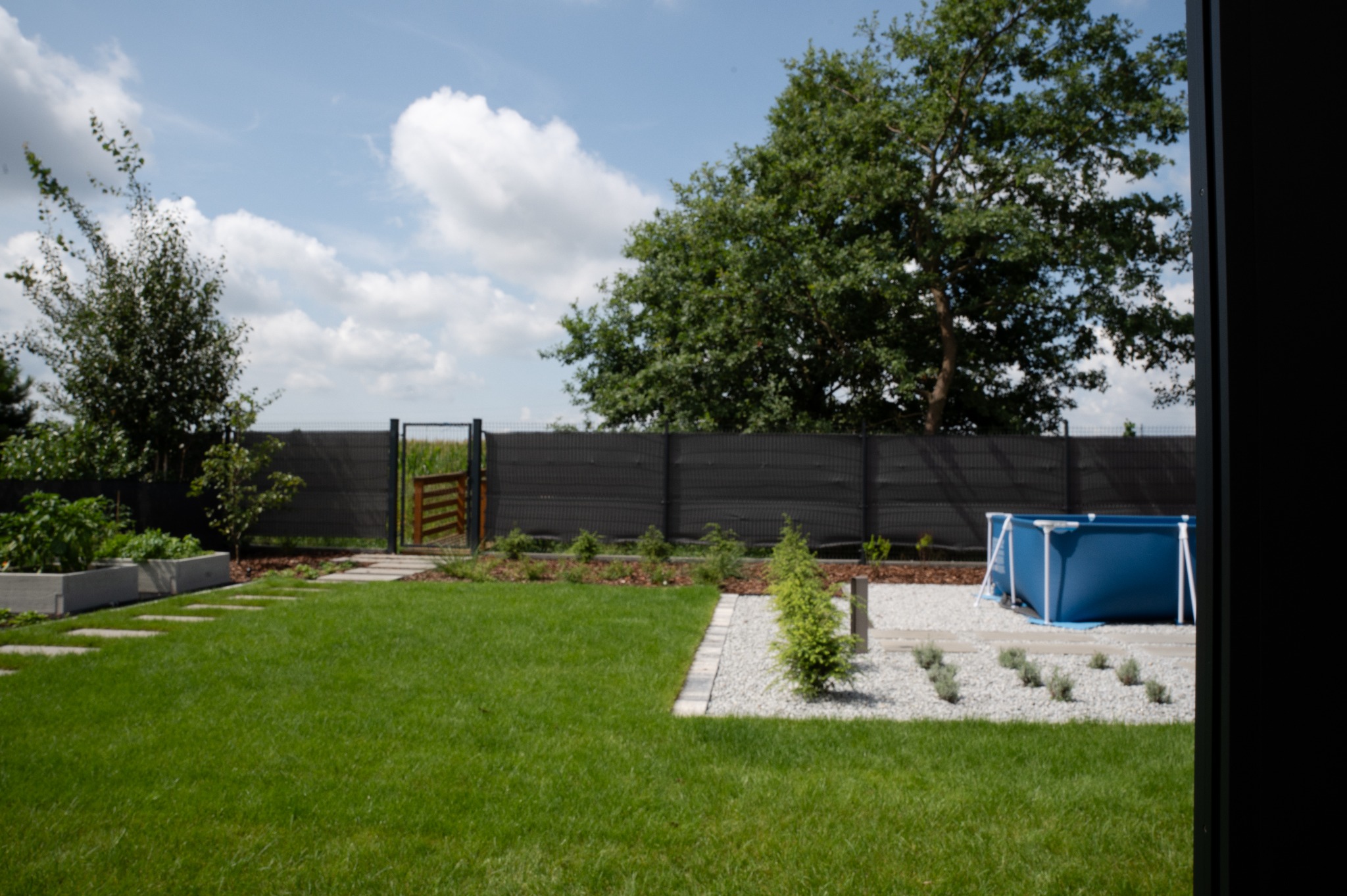 Zadbany ogród z równo przystrzyżonym trawnikiem, ścieżką z płyt, rabatami i basenem. W tle siatka ogrodzeniowa i brama. Widok zza okna.