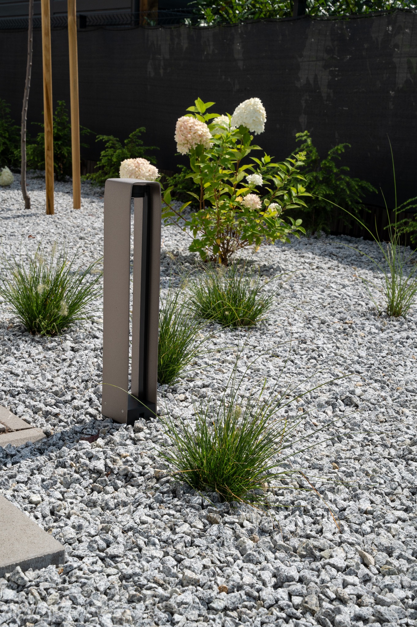 Nowoczesny ogród z białym żwirem, ozdobnymi trawami i lampą ogrodową. W tle kwitnący krzew hortensji i drewniane pale. Minimalistyczny design.