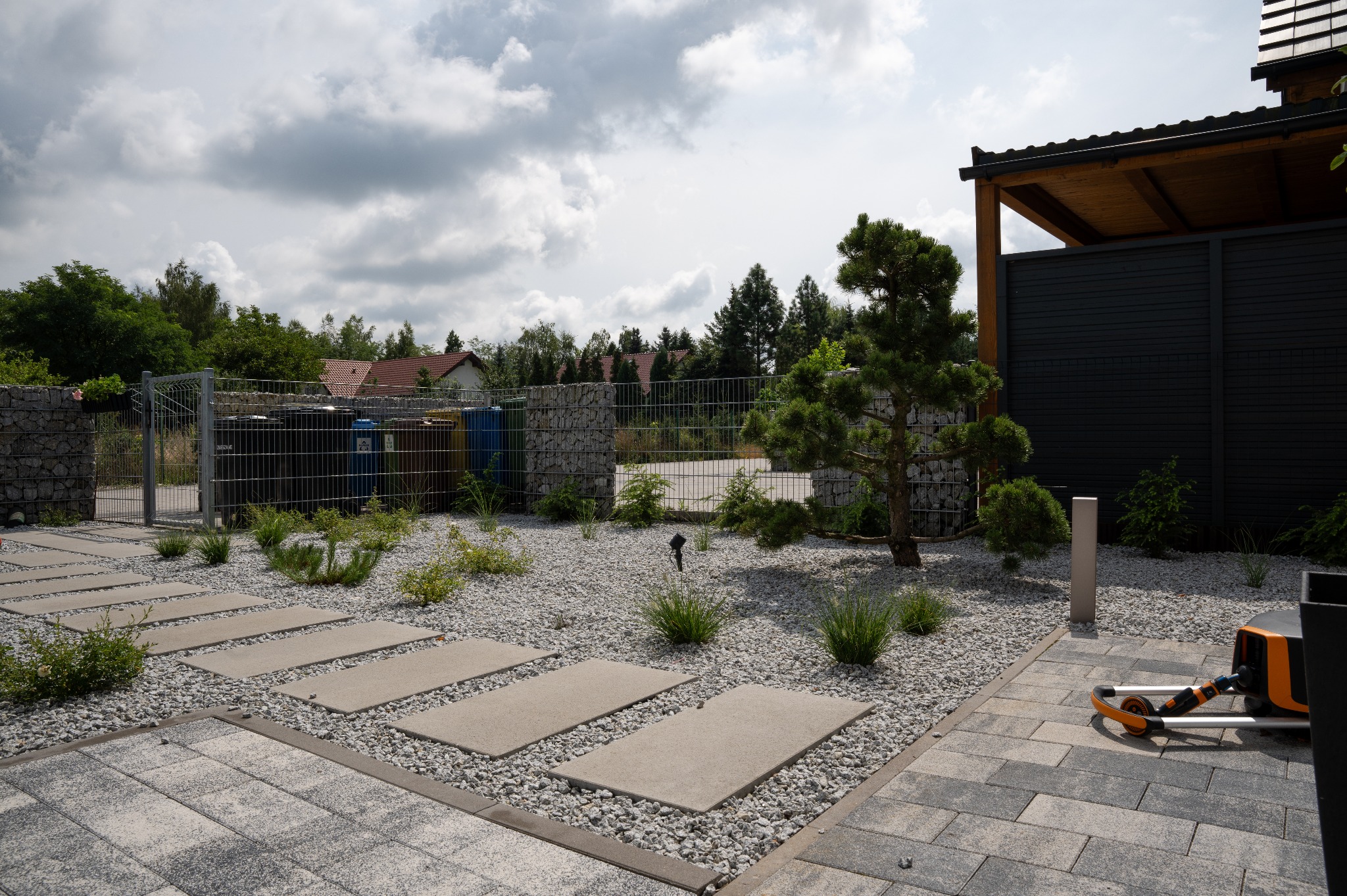 Nowoczesny ogród z ścieżką z płyt betonowych, wysypany białym żwirem, z ozdobnymi krzewami i drzewkiem bonsai. W tle ogrodzenie z gabionów i czarny płot.