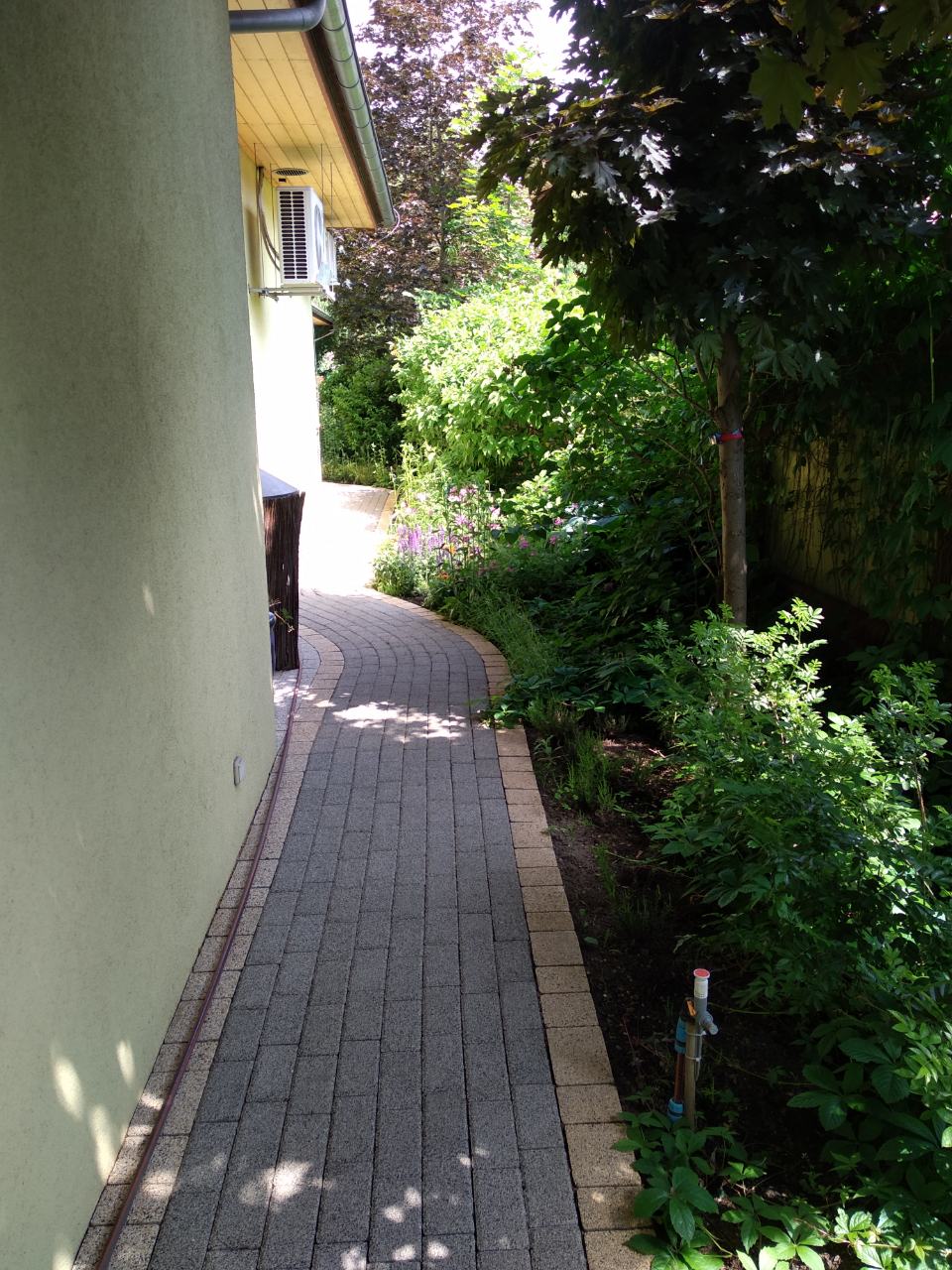 Kręta ścieżka z kostki brukowej prowadząca wzdłuż ściany budynku, otoczona bujną zielenią i drzewami, z widocznym kranem ogrodowym i klimatyzatorem na ścianie.