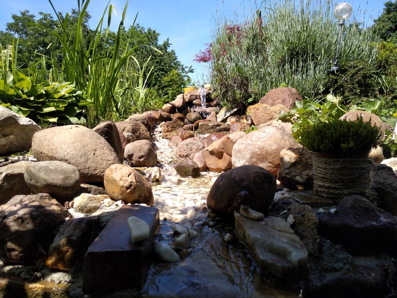 Ogród skalny z kaskadą wodną, otoczony zielenią i roślinami, z dekoracyjną lampą solarną w tle.