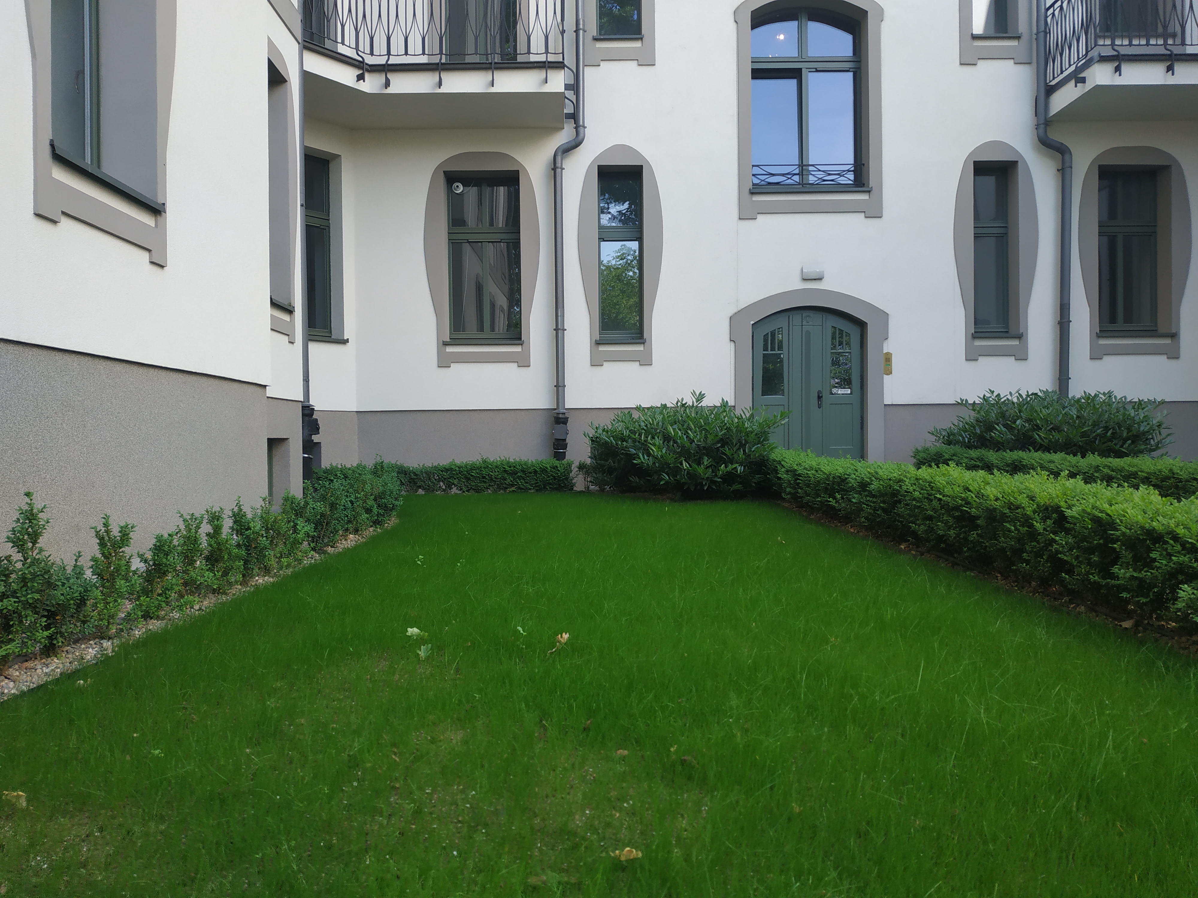 Zadbany trawnik przed budynkiem z ozdobnymi krzewami i zielonymi drzwiami wejściowymi.