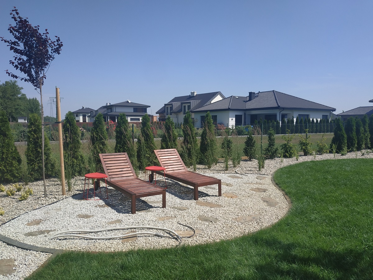 Uporządkowany ogród przydomowy z dwoma drewnianymi leżakami i czerwonymi stolikami na żwirowym podłożu, otoczony niskimi tujami i zielonym trawnikiem, z nowoczesnymi domami w tle.