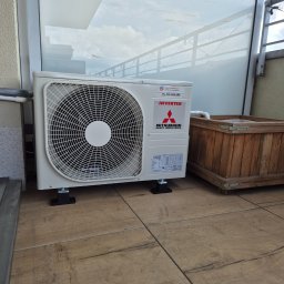 Klima-Premium Tomasz Bronowicz - Biała jednostka zewnętrzna klimatyzacji Mitsubishi Inverter na balkonie z drewnianą donicą, widok z boku, na płytkach imitujących drewno, z widocznym numerem telefonu.