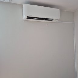 Klima-Premium Tomasz Bronowicz - Biały klimatyzator Mitsubishi Electric zamontowany na szarej ścianie w minimalistycznym wnętrzu. Widoczne przewody i gniazdko elektryczne.