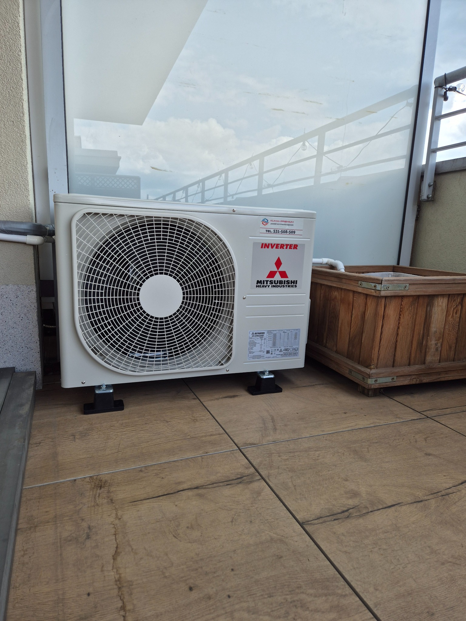 Biała jednostka zewnętrzna klimatyzacji Mitsubishi Inverter na balkonie z drewnianą donicą, widok z boku, na płytkach imitujących drewno, z widocznym numerem telefonu.