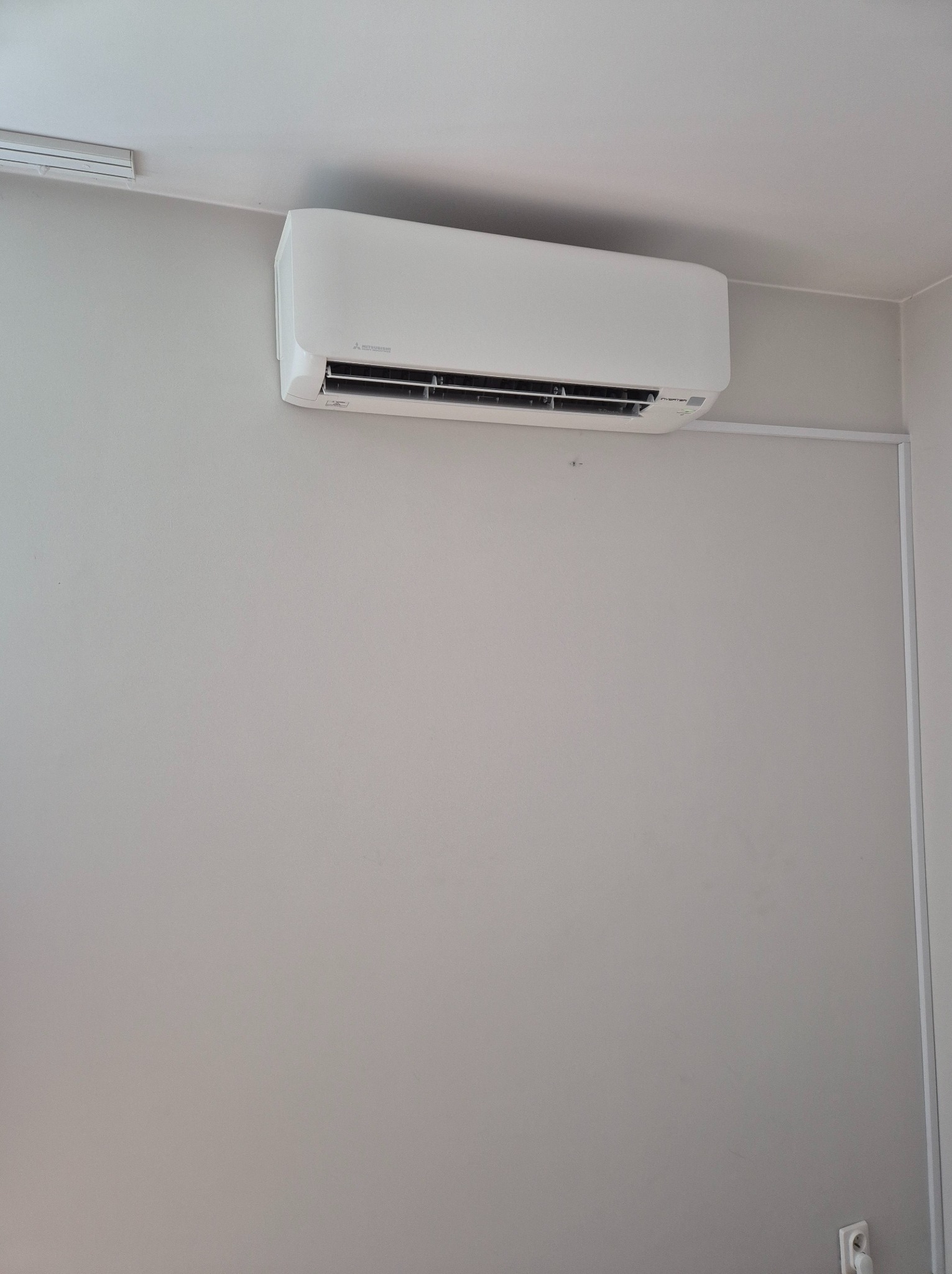 Biały klimatyzator Mitsubishi Electric zamontowany na szarej ścianie w minimalistycznym wnętrzu. Widoczne przewody i gniazdko elektryczne.
