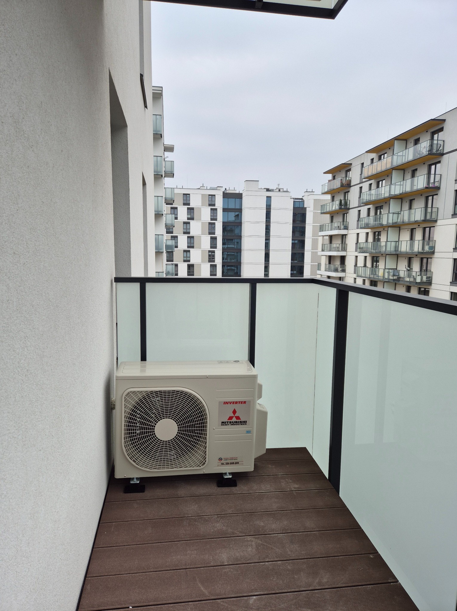 Klimatyzator Mitsubishi na balkonie nowoczesnego apartamentowca. Widok na sąsiednie budynki. Montaż klimatyzacji w przestrzeni miejskiej.