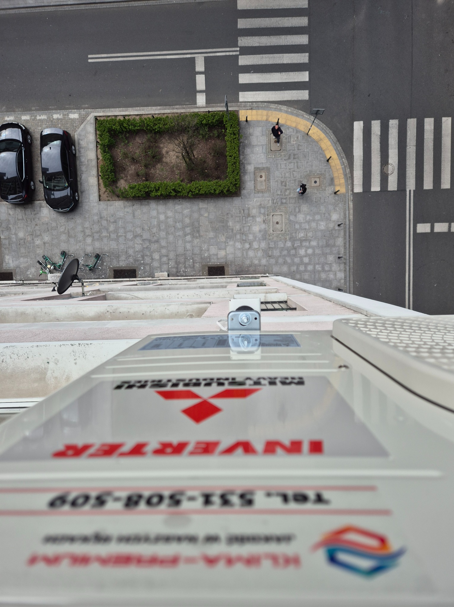 Zewnętrzna jednostka klimatyzacji Mitsubishi Inverter na tle miejskiego krajobrazu z perspektywy z góry. Widoczny fragment ulicy z przejściem dla pieszych i zaparkowanymi autami.