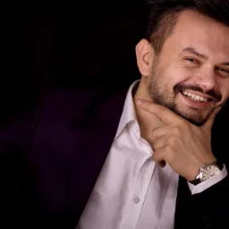 Uśmiechnięty mężczyzna w garniturze i białej koszuli, z zegarkiem na ręce, pozuje na czarnym tle, delikatnie dotykając brody palcem.