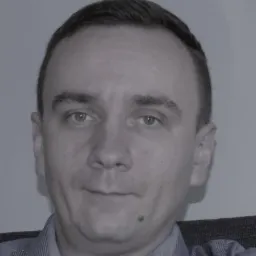 Portret mężczyzny w koszuli w paski, z pieprzykiem na brodzie, na szarym tle.