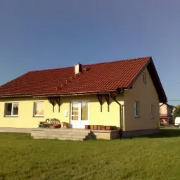 Żółty dom parterowy z czerwoną dachówką, widok z trawnika, na schodach przed wejściem doniczki i drewniana ławka.