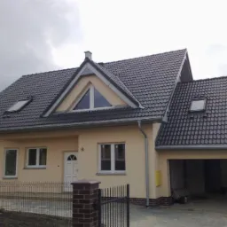 Gotowy dom jednorodzinny z szarą dachówką, beżową elewacją i garażem w bryle budynku, widoczny zza czarnego, metalowego ogrodzenia.