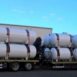 Transport białych zbiorników na gaz opałowy zabezpieczonych pasami na naczepie ciężarówki, widoczne opony i fragment budynku w tle.