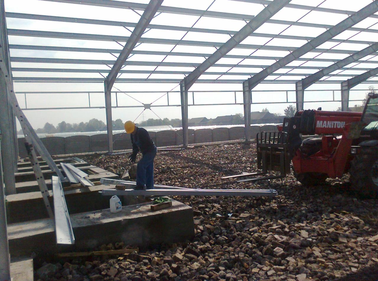 Pracownik w żółtym kasku przy montażu stalowej konstrukcji hali. Na pierwszym planie drabina i elementy konstrukcyjne, w tle widoczny podnośnik teleskopowy Manitou oraz krajobraz.