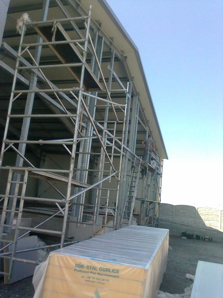 Konstrukcja stalowa hali w trakcie montażu, widoczne rusztowania i pracownik na drabinie, w tle składowane płyty warstwowe z widocznym logo producenta.
