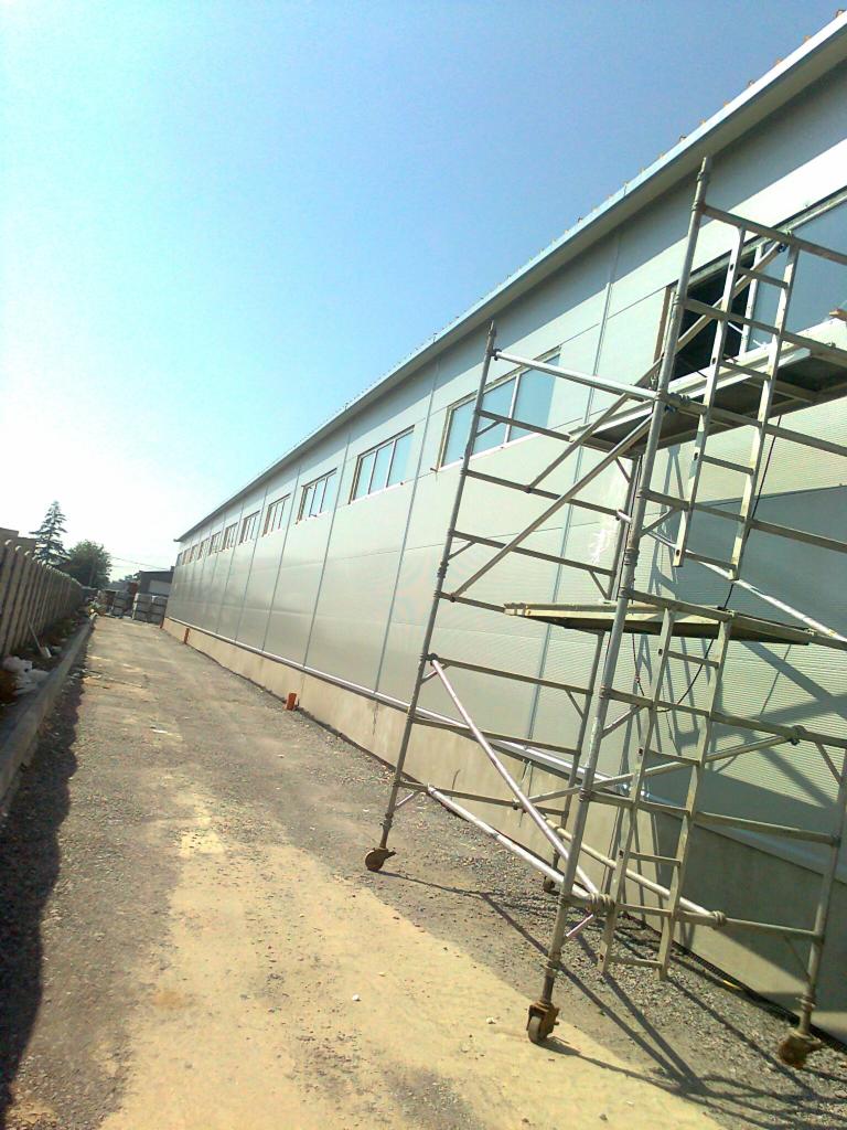 Długa hala stalowa z szarej płyty warstwowej, widoczna z boku, z rusztowaniem ustawionym wzdłuż ściany, wskazującym na trwający montaż.