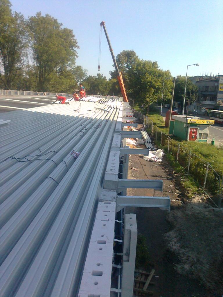 Widok z góry na montaż dachu z płyt warstwowych na konstrukcji stalowej hali, z dźwigiem w tle i pracownikami w czerwonych kombinezonach.