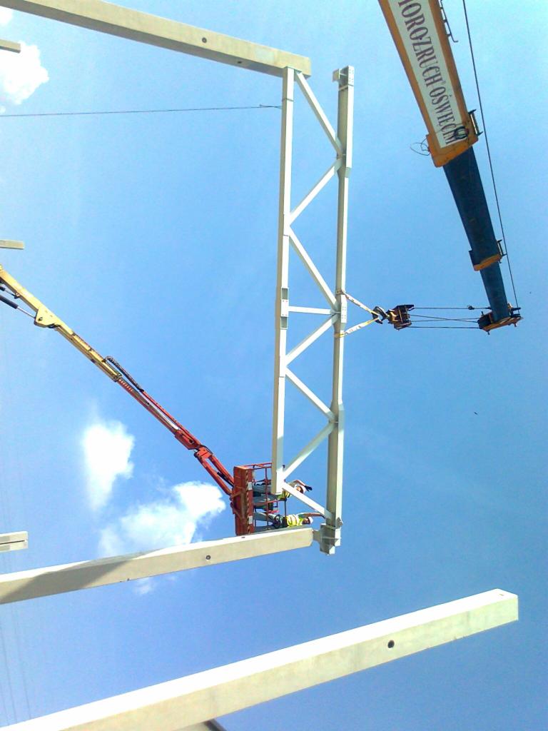 Montaż białej konstrukcji stalowej hali z użyciem dźwigu i podnośnika koszowego na tle błękitnego nieba.