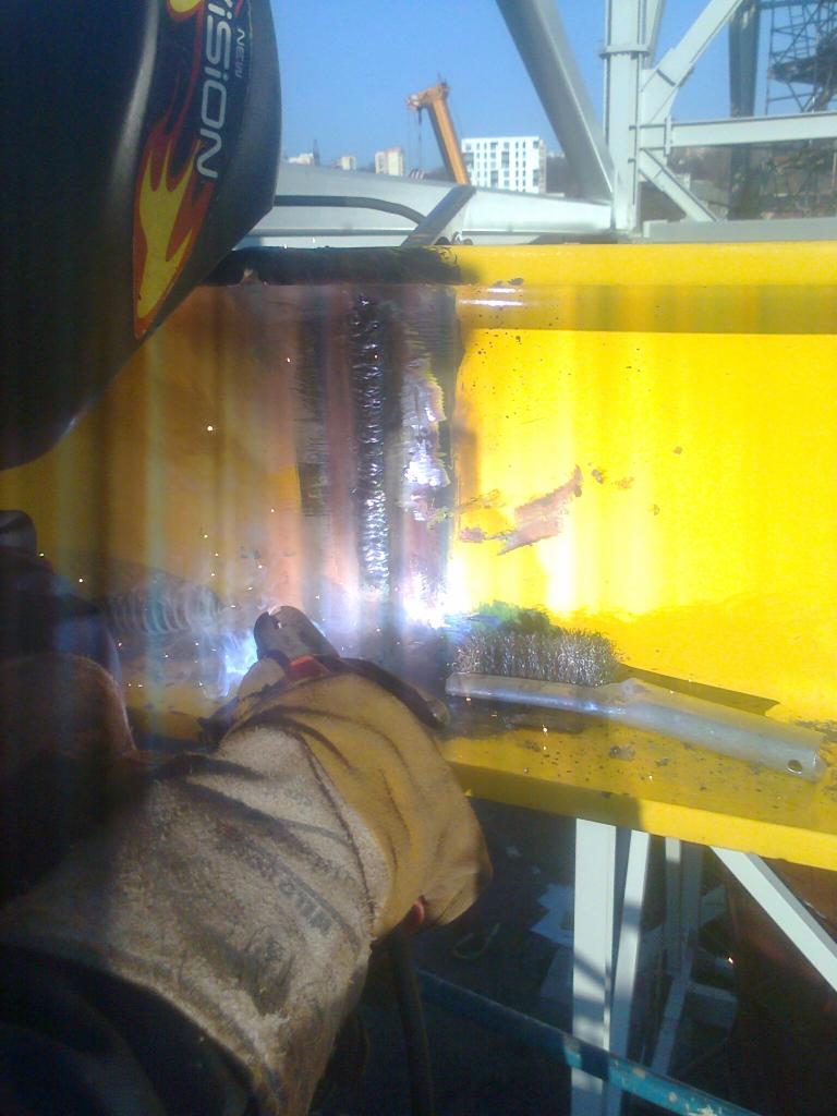 Spawanie żółtej, stalowej belki konstrukcyjnej na budowie, widoczne iskry, spawacz w masce i rękawicach ochronnych, obok szczotka druciana do czyszczenia spoin, w tle budynki i dźwig.