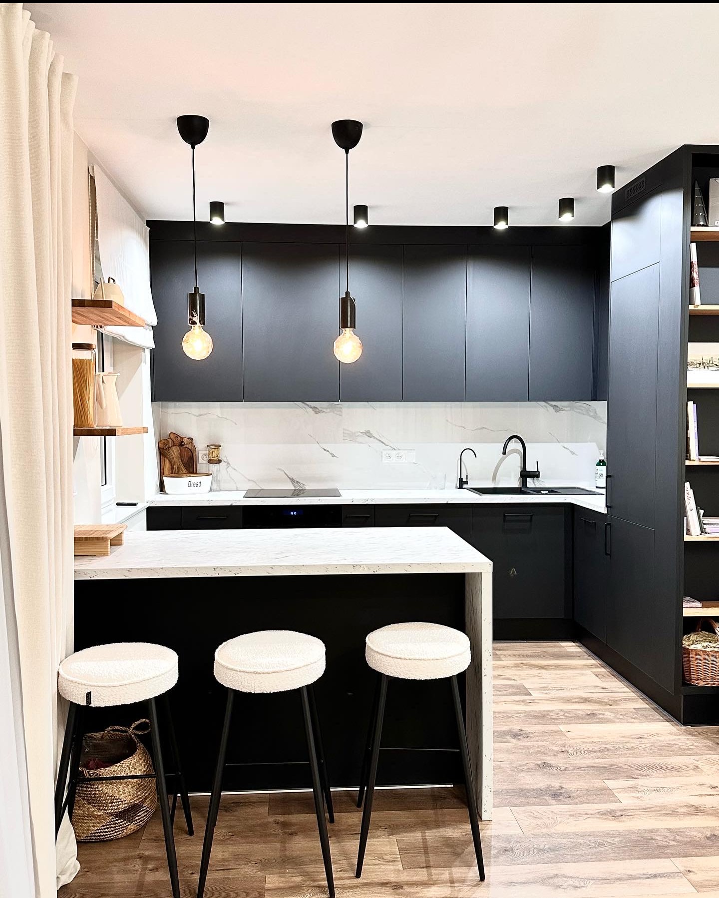 Nowoczesna kuchnia w odcieniach czerni i bieli z marmurowym blatem, oświetlona wiszącymi lampami i punktowymi reflektorami, uzupełniona trzema białymi hokerami i drewnianą podłogą.