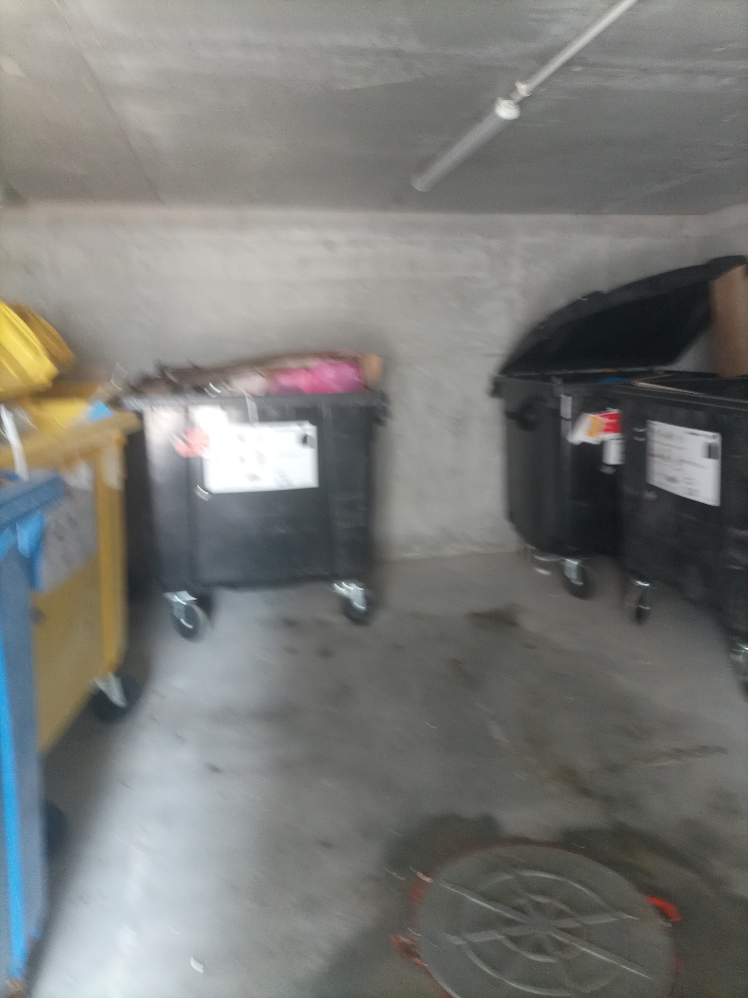 Wnętrze betonowego pomieszczenia z kilkoma pojemnikami na śmieci, w tym żółtym i czarnym, oraz okrągłą studzienką kanalizacyjną w podłodze.