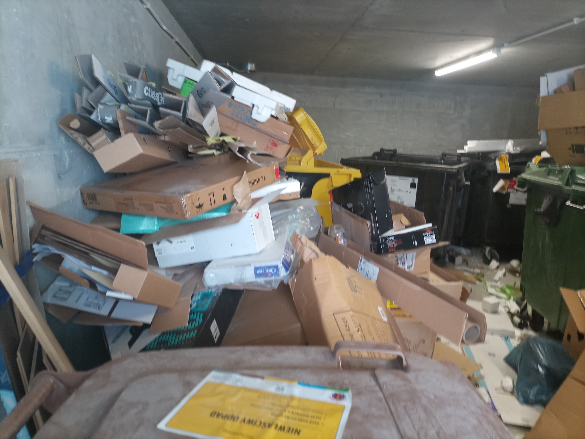 Stos kartonowych pudeł i innych śmieci w pomieszczeniu magazynowym, przy ścianie i obok kontenerów na śmieci.