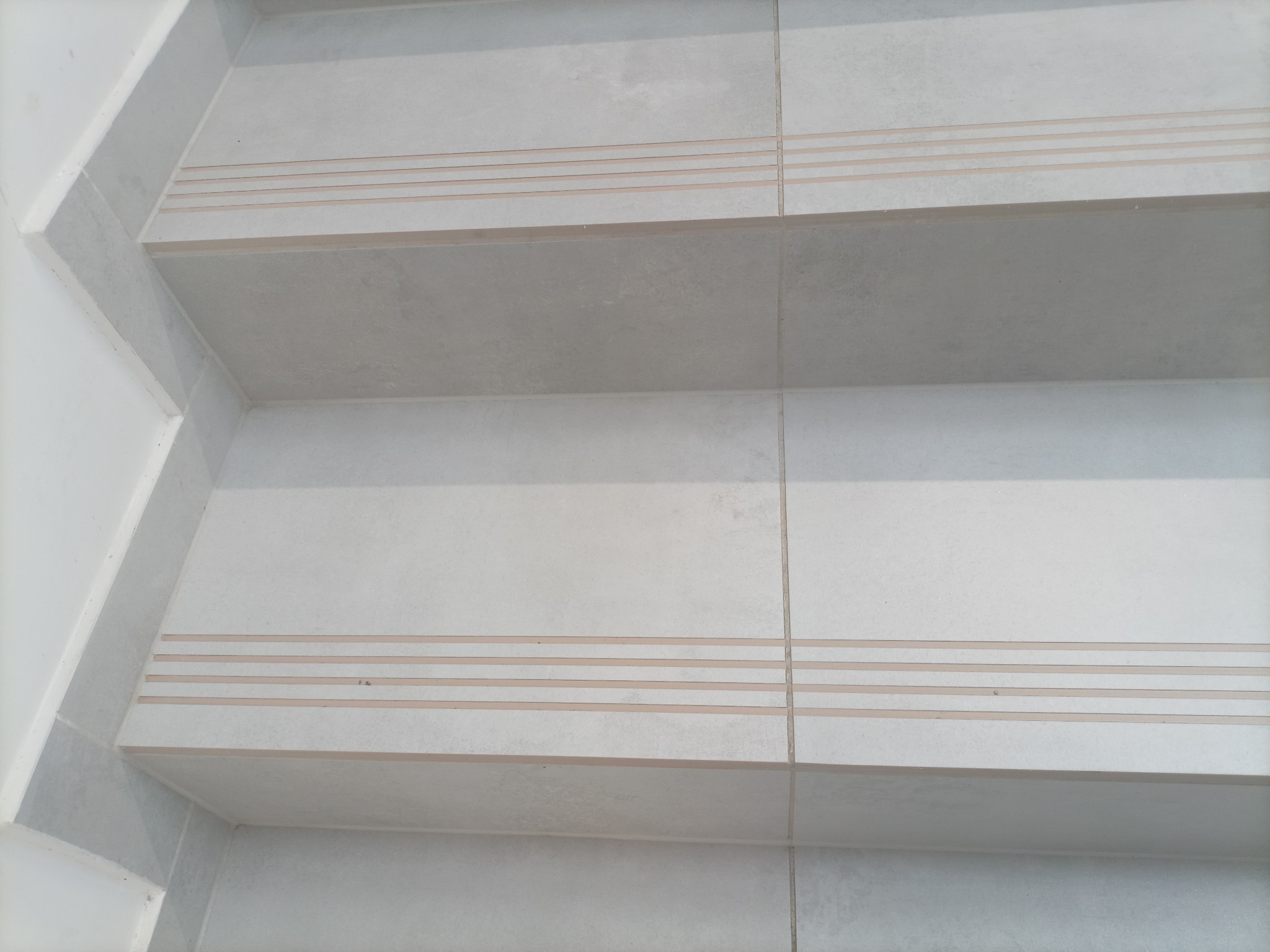 Jasnoszare płytki ceramiczne na schodach wewnętrznych z trzema poziomymi paskami antypoślizgowymi na krawędziach stopni.