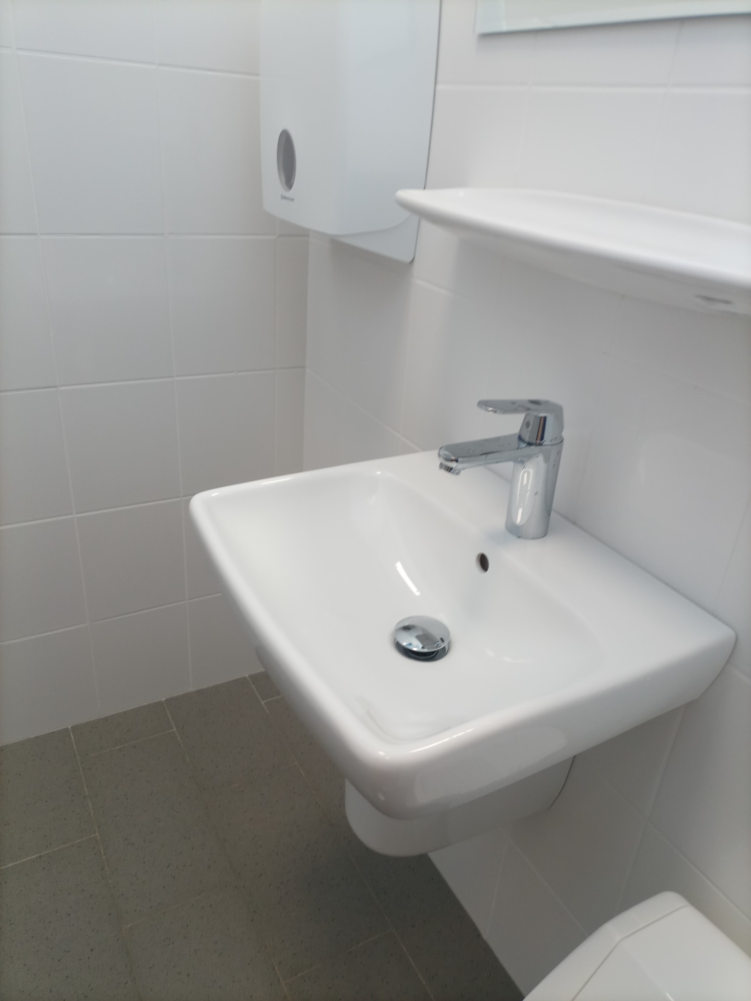 Biała umywalka z chromowaną baterią i odpływem w czystej, wykafelkowanej łazience z dozownikiem na ręczniki.