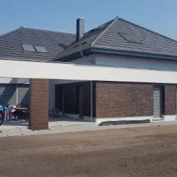 Nowoczesny dom jednorodzinny z grafitowym dachem i elewacją w kolorze złamanej bieli, z elementami dekoracyjnymi imitującymi drewno. Teren wokół domu przygotowany pod trawnik.