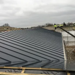 Montaż blachy na dachu budynku w trakcie budowy, widoczny dźwig i pracownik w żółtej kamizelce.