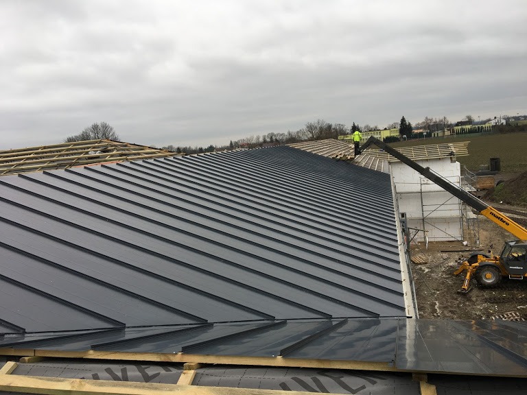 Montaż blachy na dachu budynku w trakcie budowy, widoczny dźwig i pracownik w żółtej kamizelce.