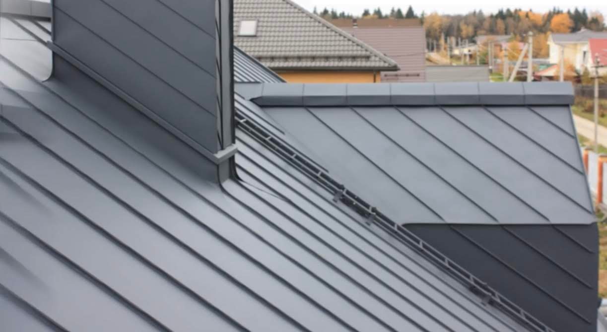 Szary dach z blachy na domu jednorodzinnym, widok z góry, z widocznym kominem i elementami wykończeniowymi, w tle zabudowa jednorodzinna i drzewa.