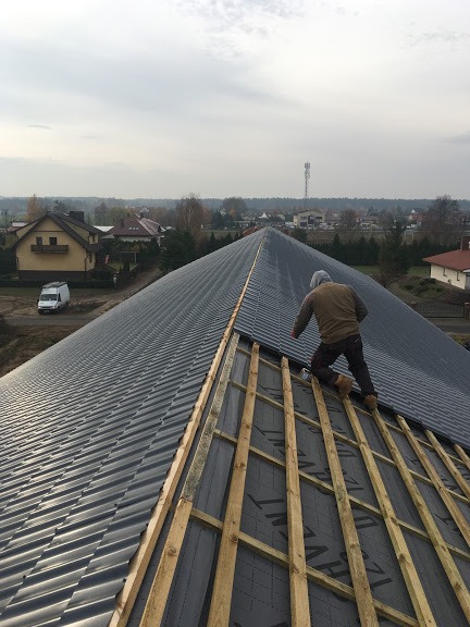 Montaż blachy na dachu szeregowca w Głogowie koło Torunia, widoczne drewniane łaty i membrana dachowa, w tle zabudowa jednorodzinna i wieża telekomunikacyjna.